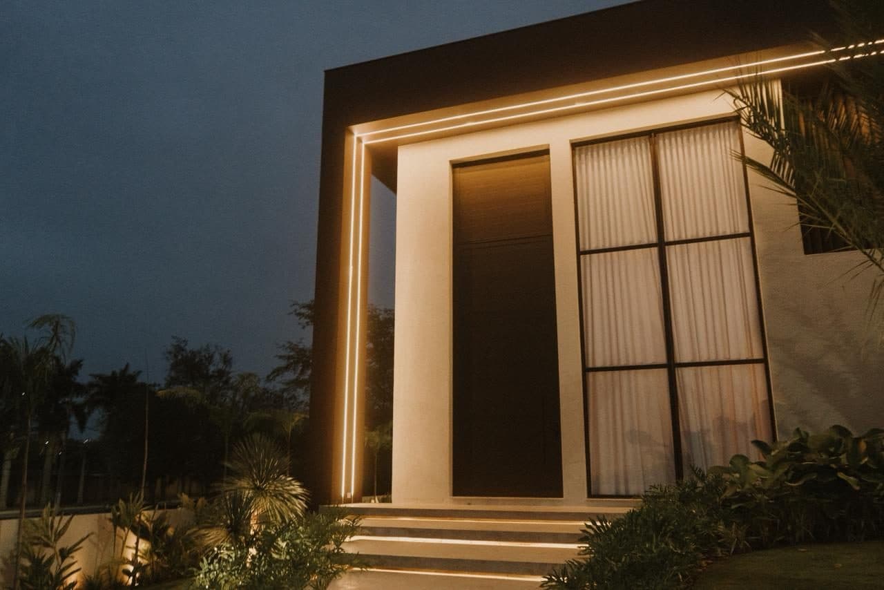 Detalhe externo noturno com entrada iluminada, portas altas e paisagismo