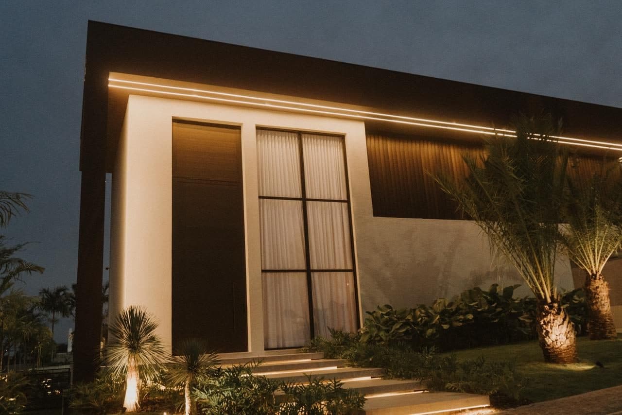 Vista externa noturna com iluminação linear na cobertura, janelas altas e jardim